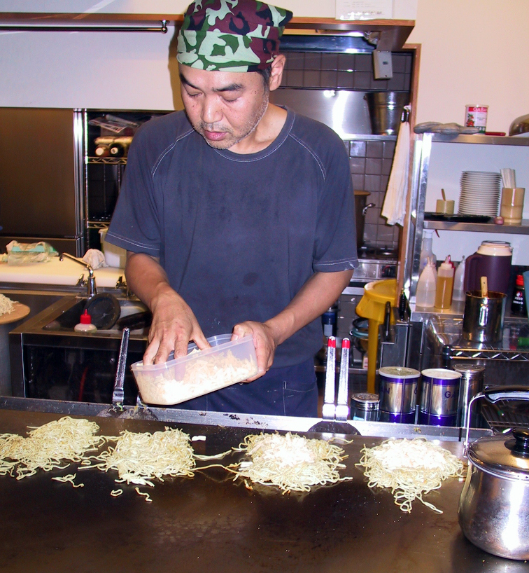 Okonomiyaki cook working at a teppan counter in Hiroshima with raw ingredients arranged for layering
