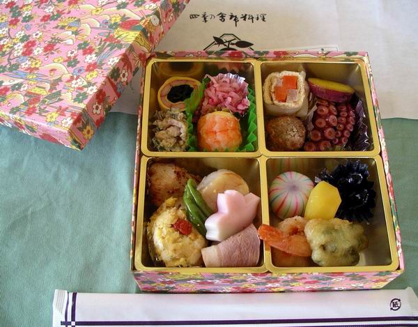 Hanami picnic with sake, cups, and a colourful bento under cherry blossoms